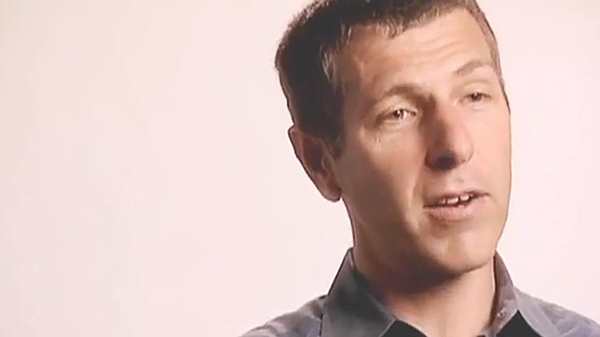 A man wearing a grey collared shirt is interviewed against a light background.