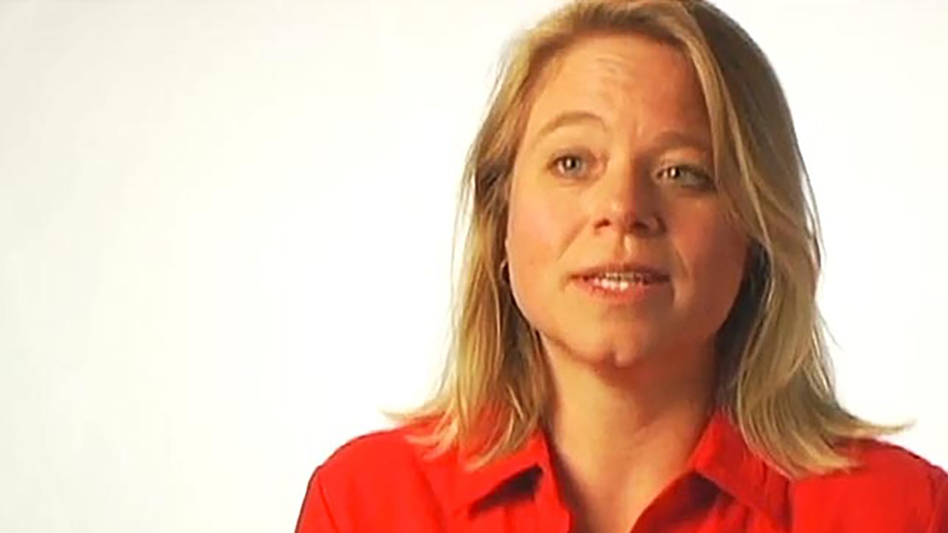 A blonde woman wearing a red shirt being interviewed against a white background