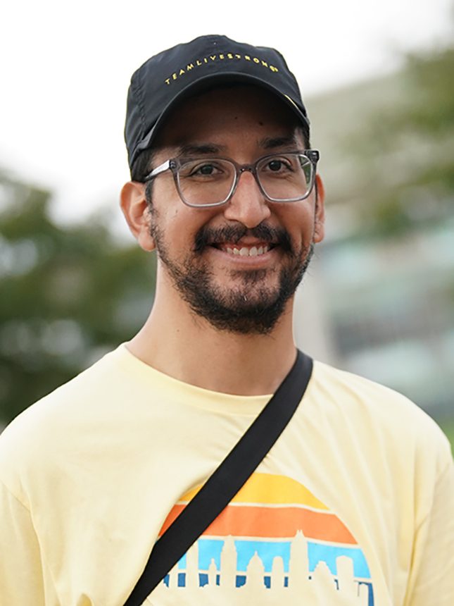 A smiling man wearing a black hat, glasses, and a yellow shirt