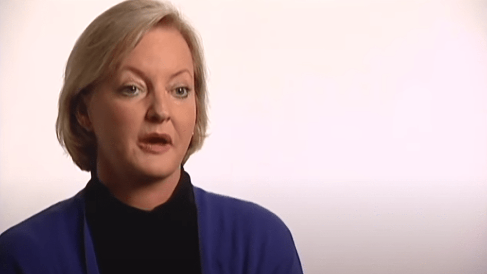 A middle-aged woman with short blonde hair wearing a black turtleneck and blue cardigan is interviewed against a white background.