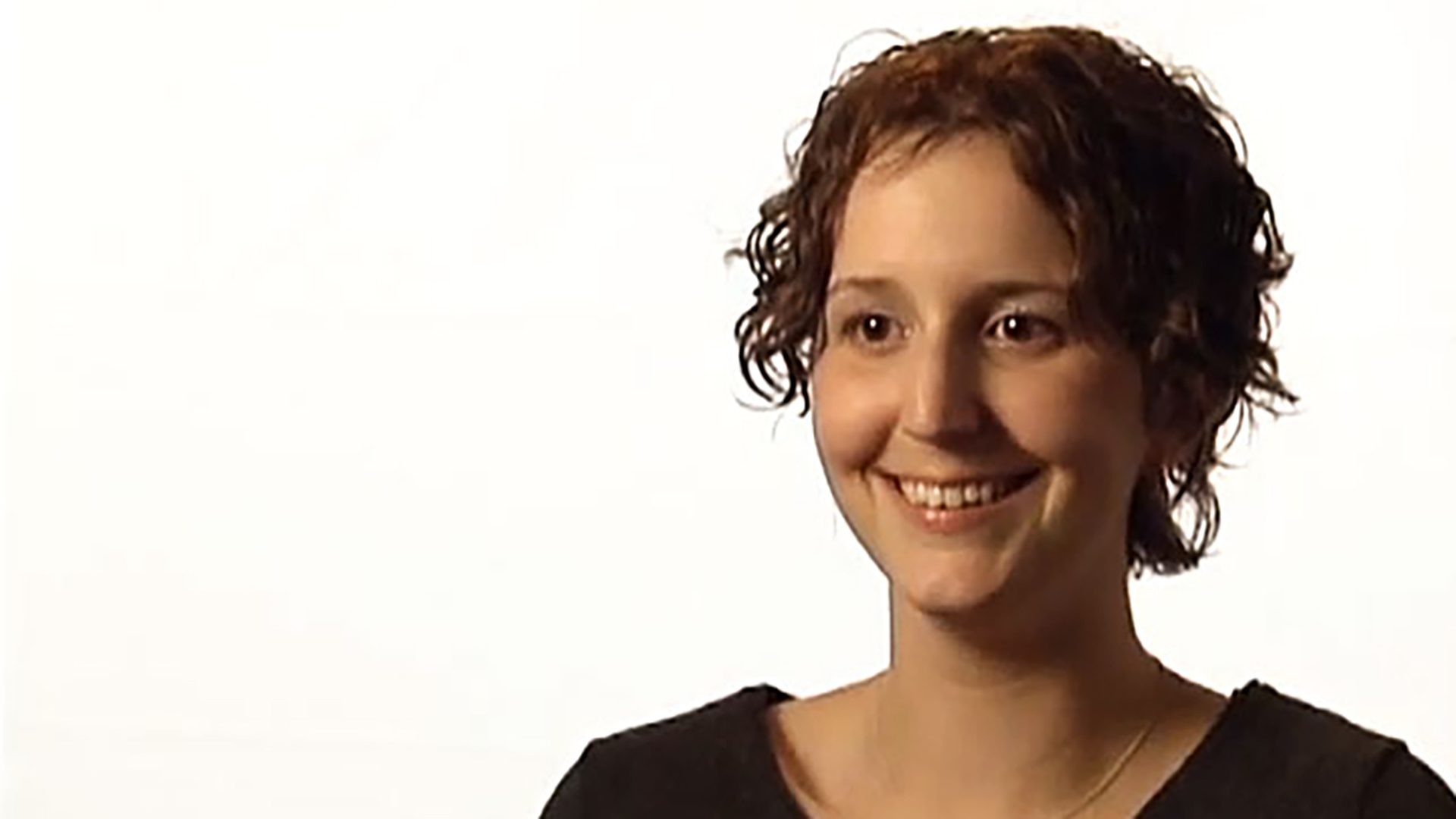 A young woman with short curly hair wears a black shirt and is interviewed against a white background.