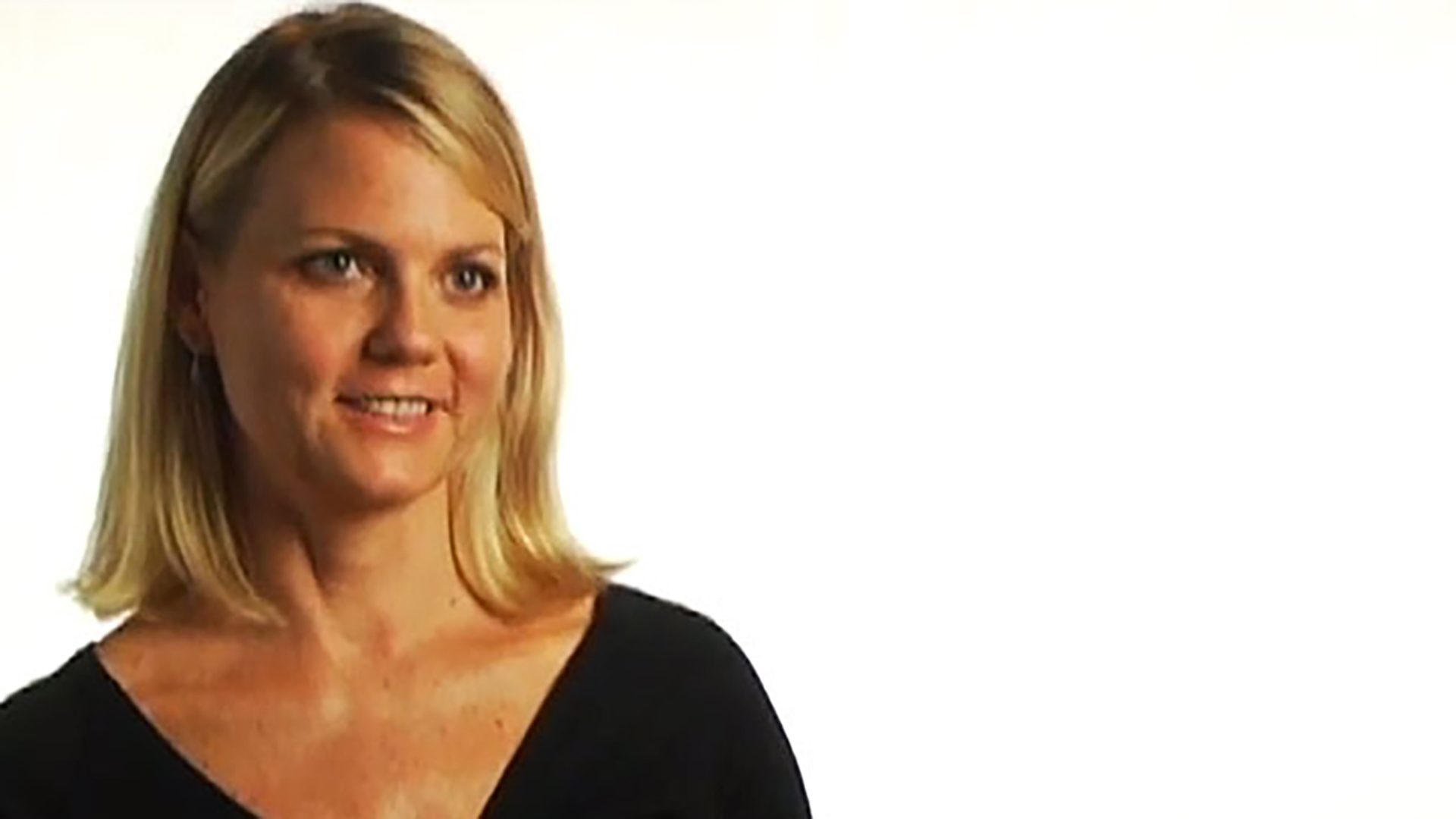 A blonde adult woman wearing a black V-neck shirt is interviewed against a white background.