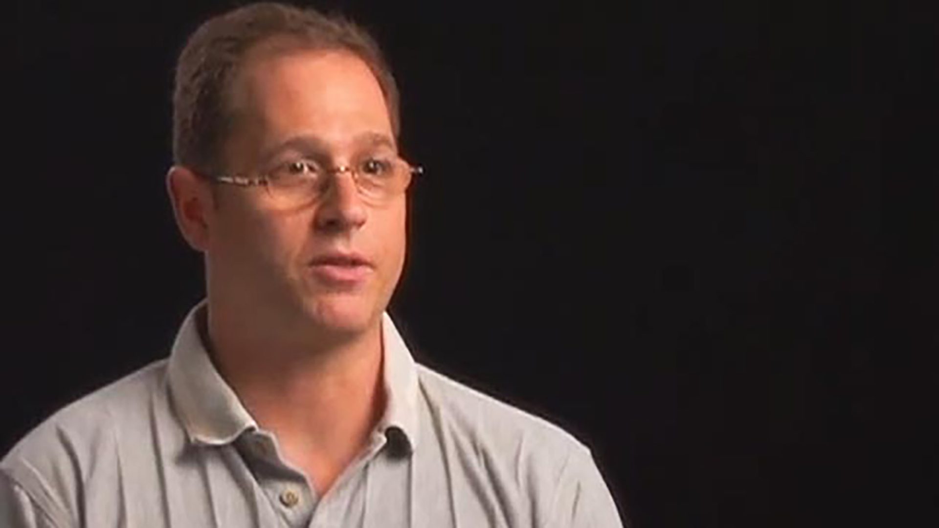 An adult man wearing glasses and a gray polo shirt is interviewed against a black background.