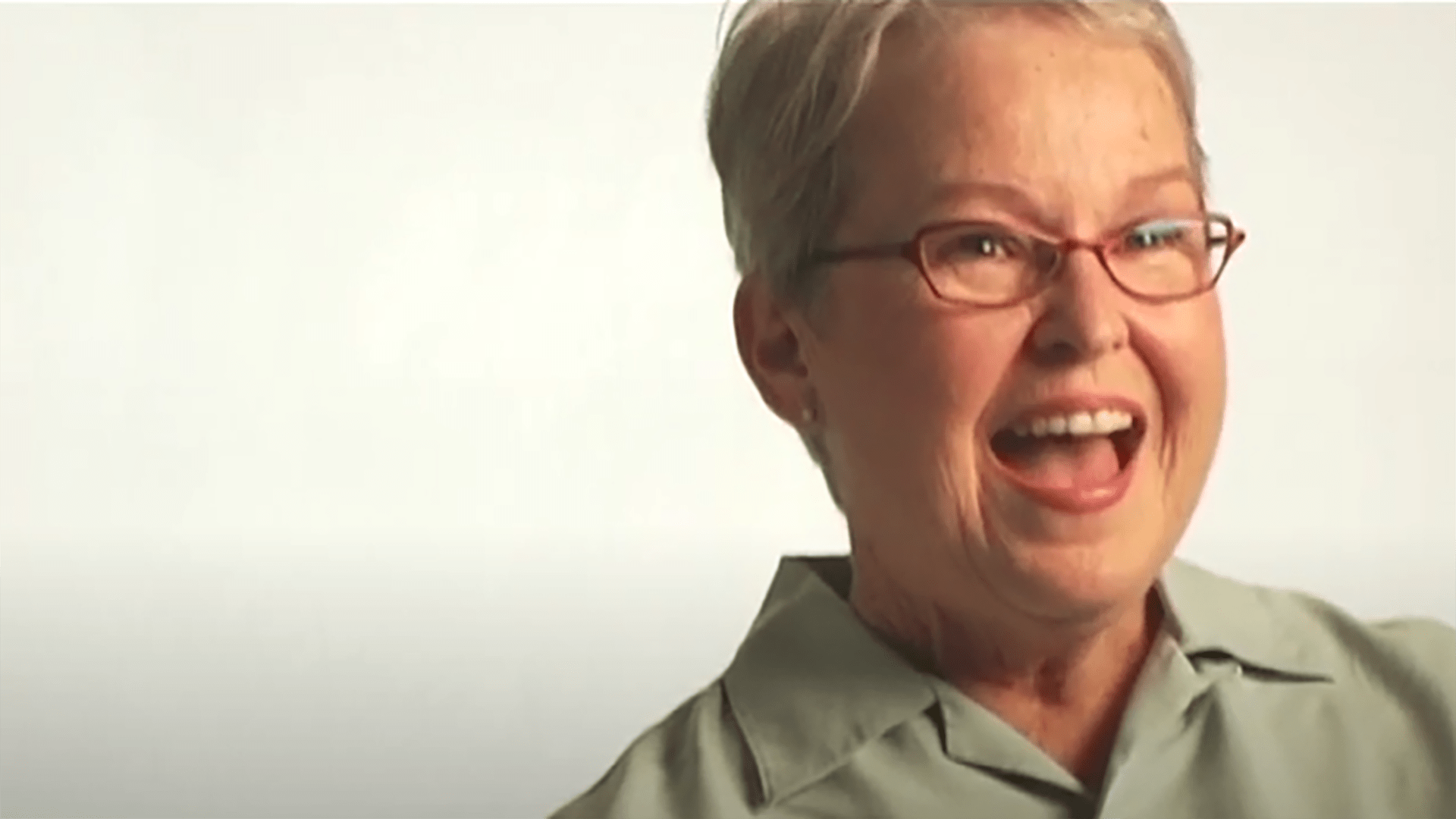 A smiling senior woman with short gray hair wearing glasses and a light green button-up shirt