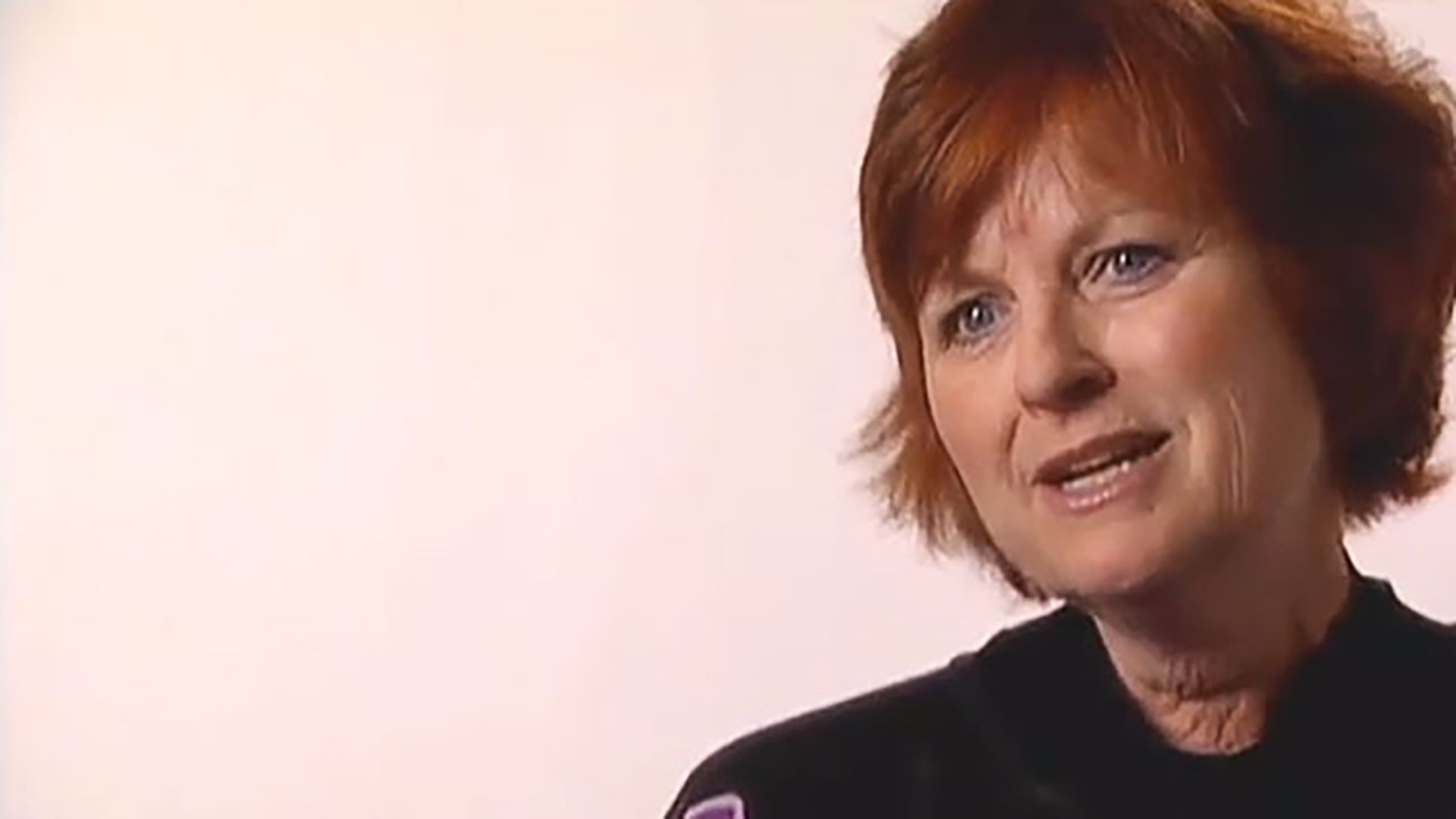A middle-aged woman with short red hair wearing a black shirt is interviewed against a light background.