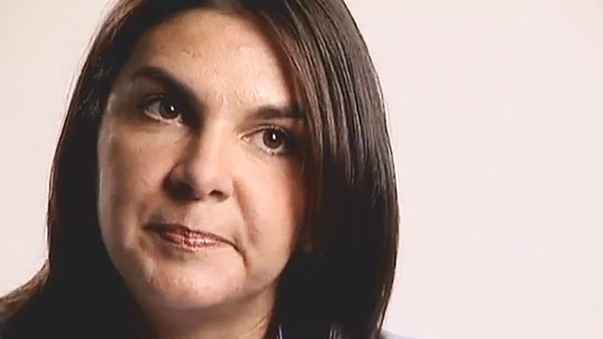 A woman with dark hair is interviewed against a light background.