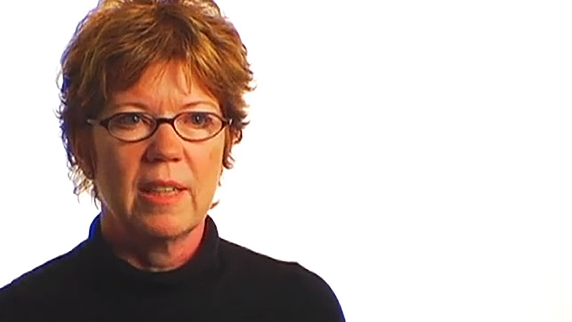 A middle-aged woman with red hair, glasses, and a black turtleneck is interviewed against a white background.