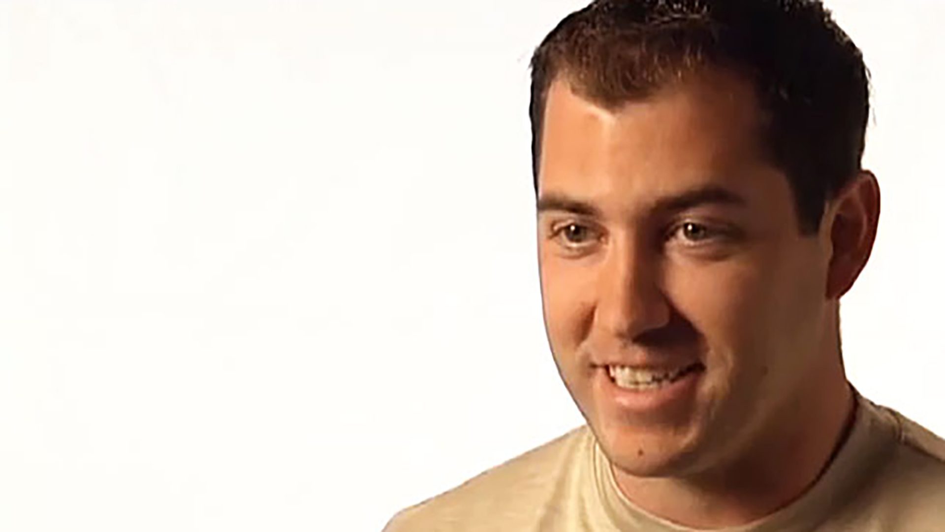 A smiling young adult man wearing a cream colored shirt is interviewed against a white background.