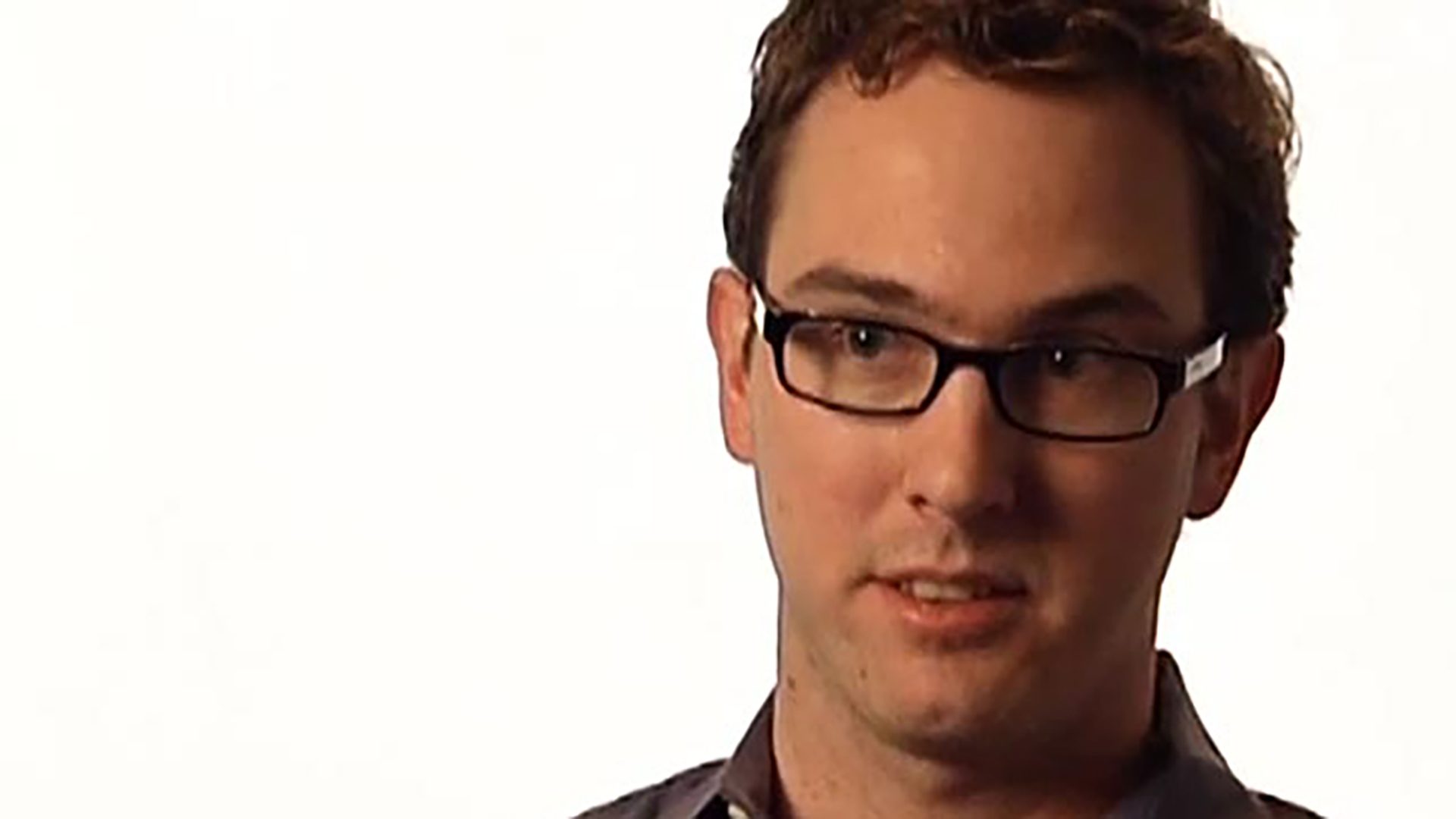A young adult man wearing black glasses is interviewed against a white background.
