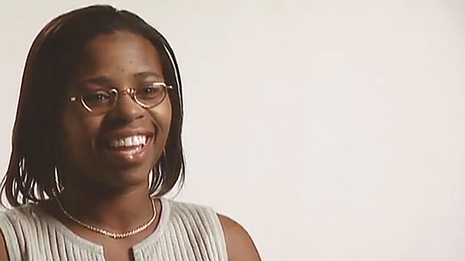 A smiling young woman wearing glasses and a gray sleeveless shirt is interviewed against a white background.