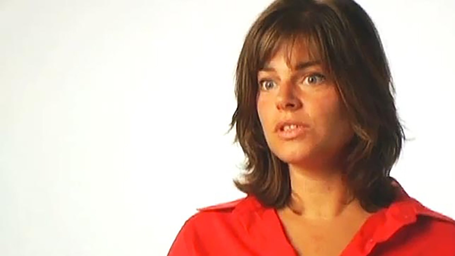 A young woman with shoulder-length brown hair wears a red shirt and is interviewed against a white background.