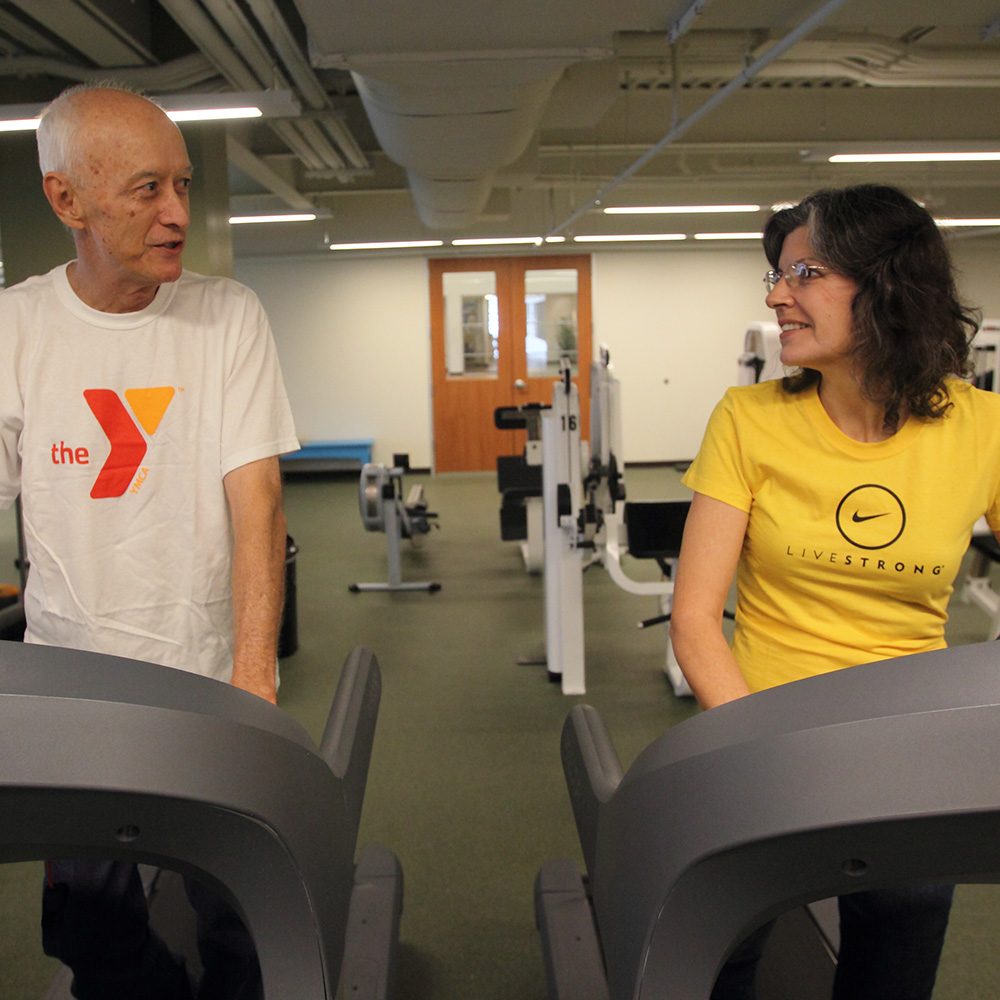 A man walking on a treadmill wearing a white YMCA shirt next to a woman walking on a treadmill wearing a yellow Livestrong shirt