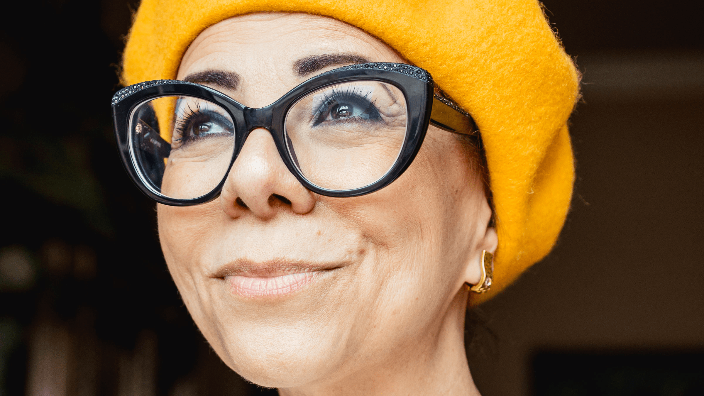 A middle aged woman wearing glasses and a yellow hat looks into the distance.