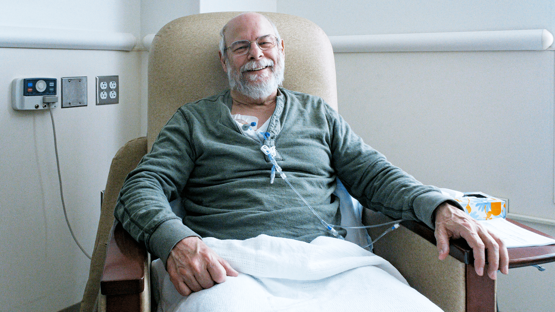 A smiling senior man sits in a chair receiving cancer treatment.