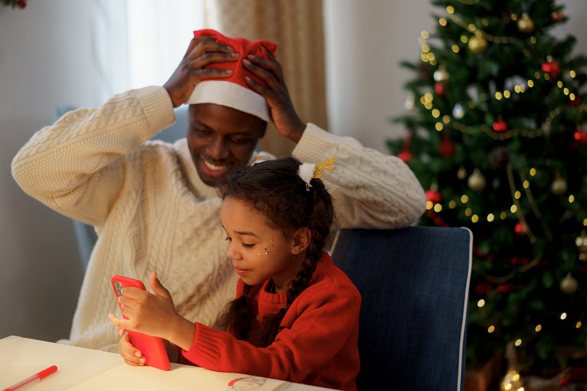 A father wearing a Santa hat supervises his young daughter using a smartphone. A Christmas tree is in the background.