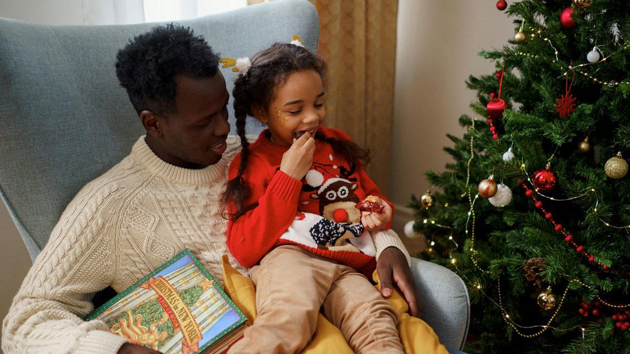 A father sits with his young daughter on his lap next to a Christmas tree