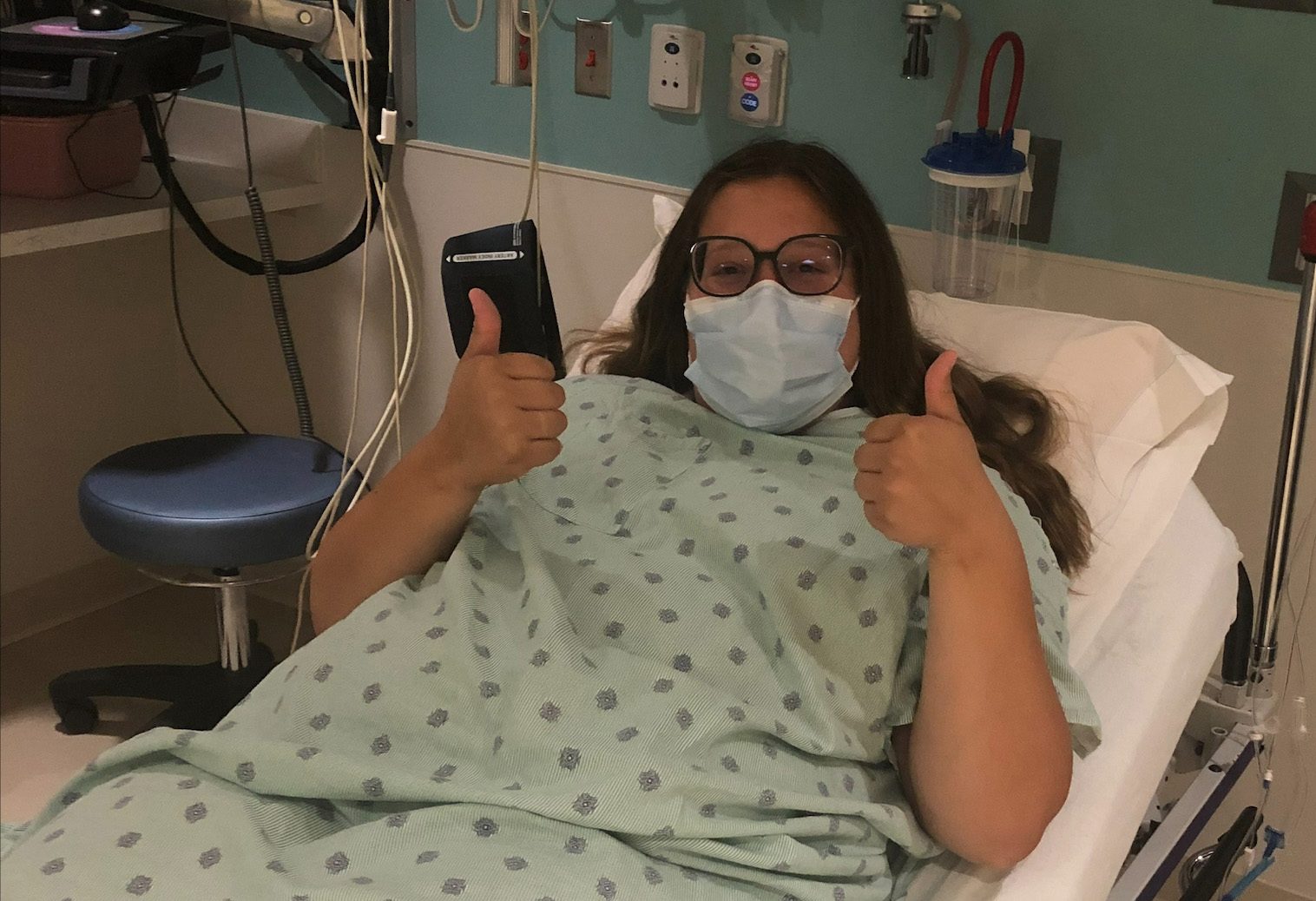 A young woman laying in a hospital bed gives 2 thumbs up