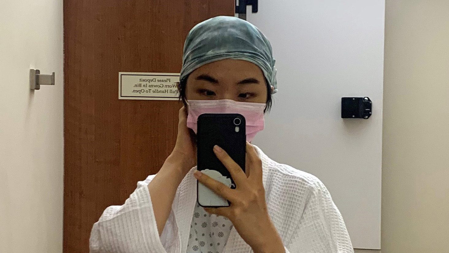 A young woman wearing a hospital gown, mask, and head wrap takes a selfie