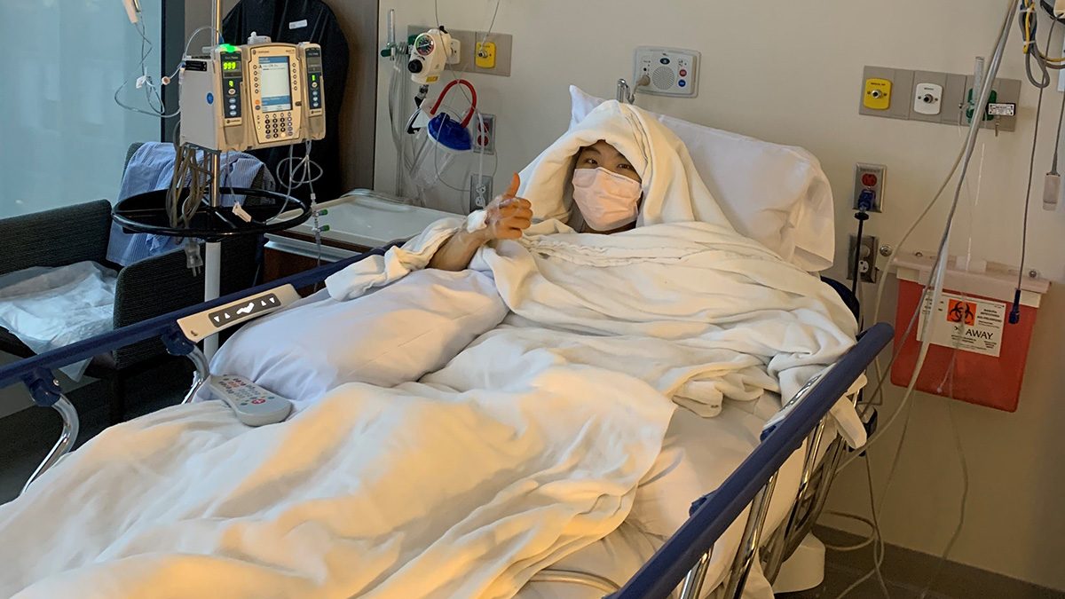 A young woman in a hospital bed gives a thumbs up