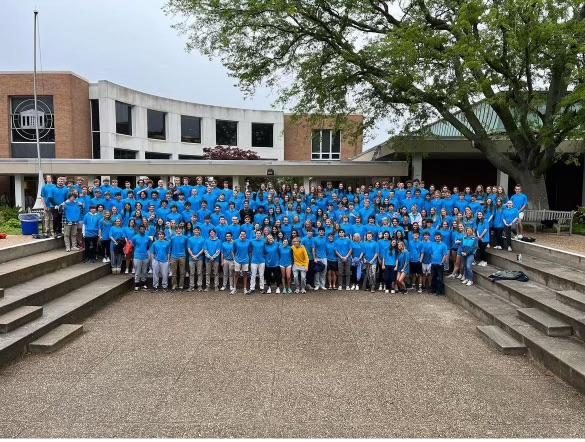 A large group of students stand in front of a school wearing matching blue shirts