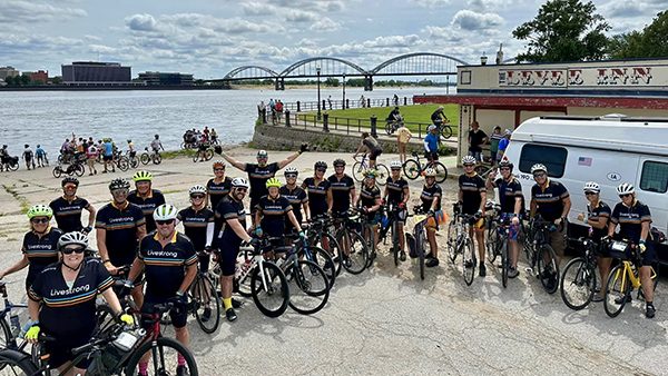 A group of cyclists wearing black jerseys poses with their bikes next to a body of water