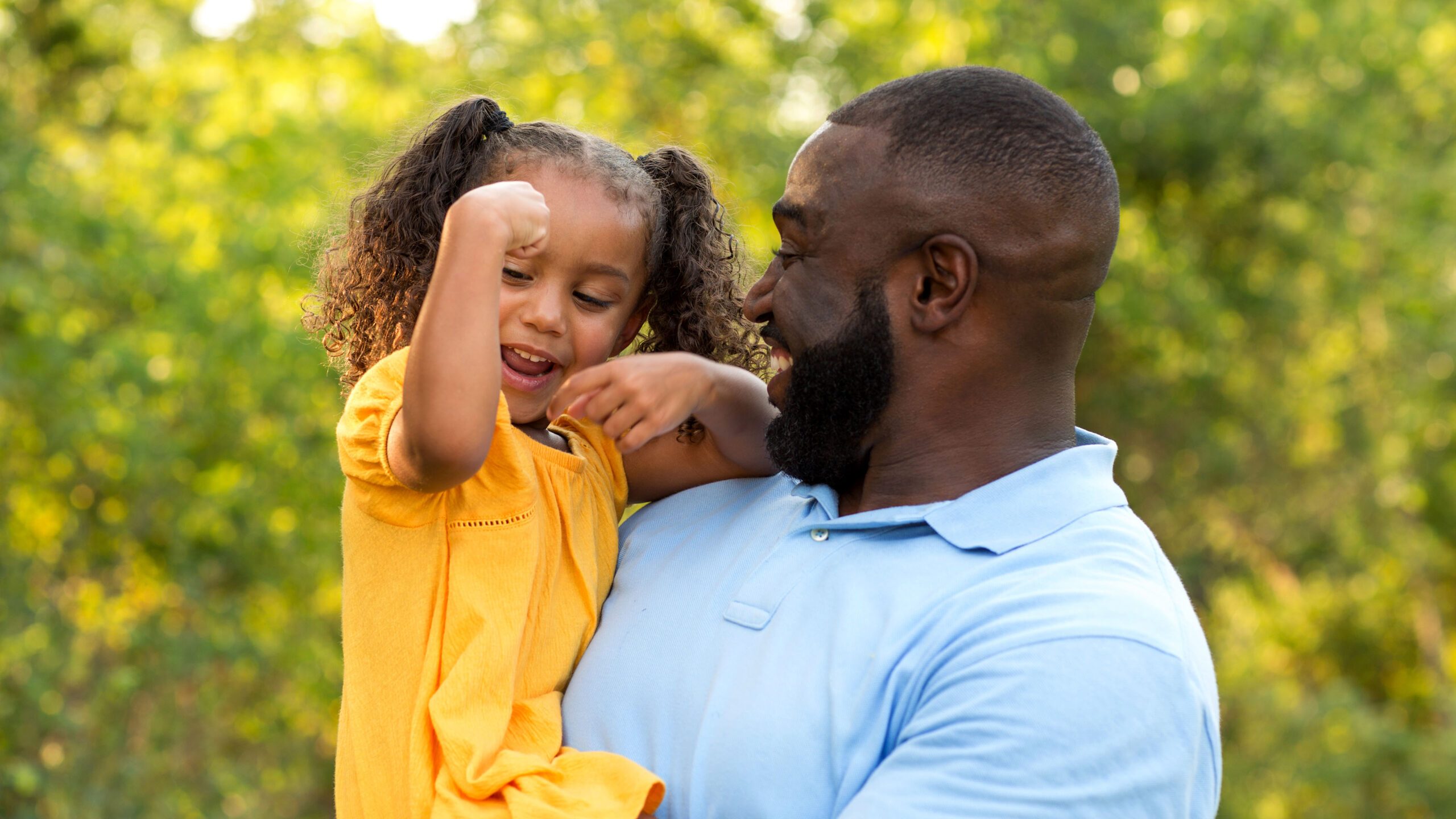 A smiling father holds his young daughter, who flexes her arm muscle.