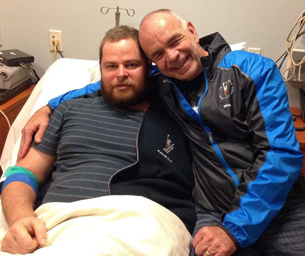 A man sits in a hospital bed while his friend embraces him