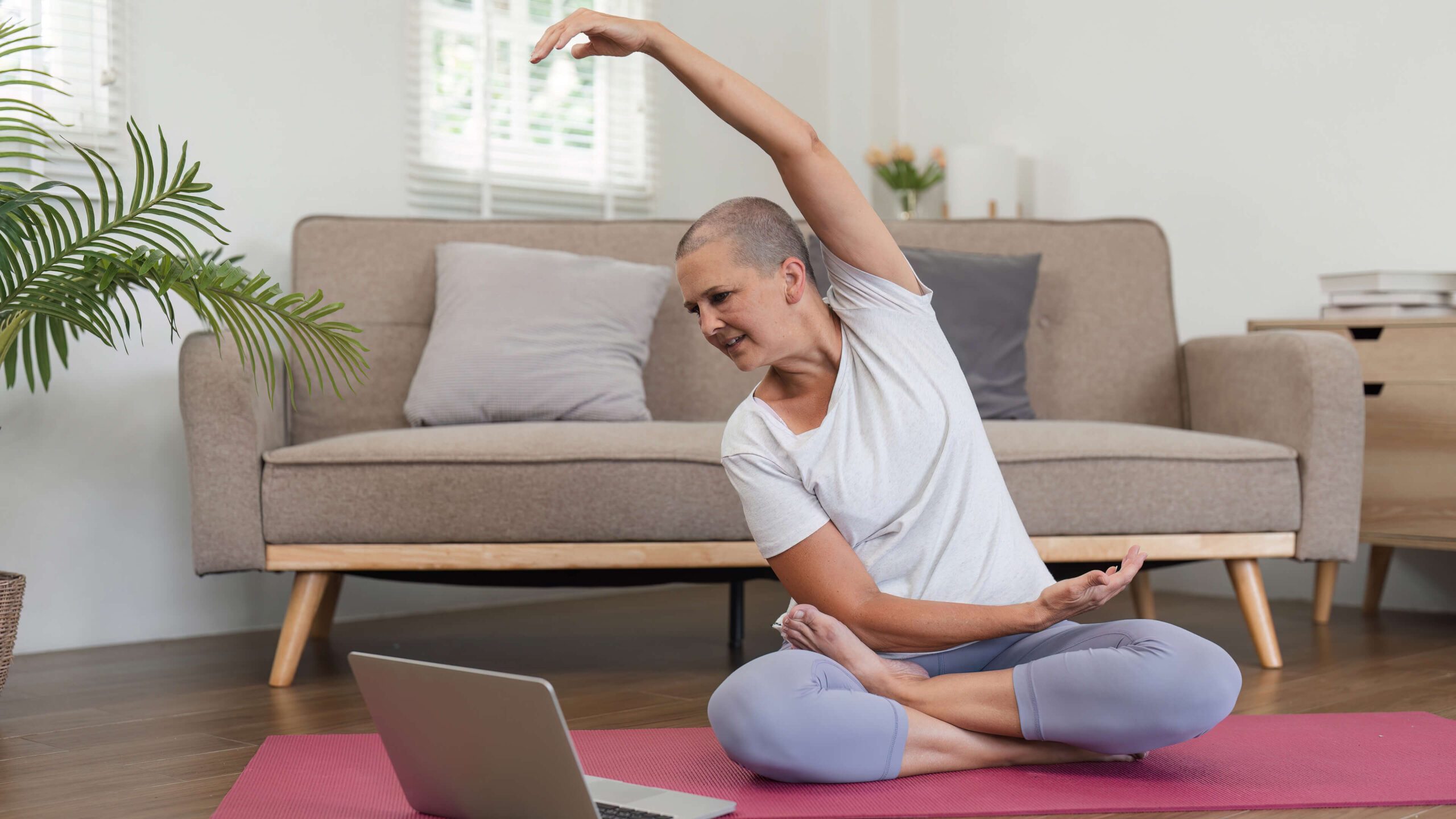 A woman sitting and stretching on a yoga mat watching a workout video on a laptop